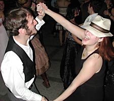 He wears a vest; she wears a man's hat. They laugh as they dance.