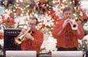 Jim Chamberlain and Brad Davies play trumpets in front of a Christmas tree.
