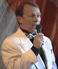 Kirk sings in a white sport coat, black shirt, and black-and-white tie.